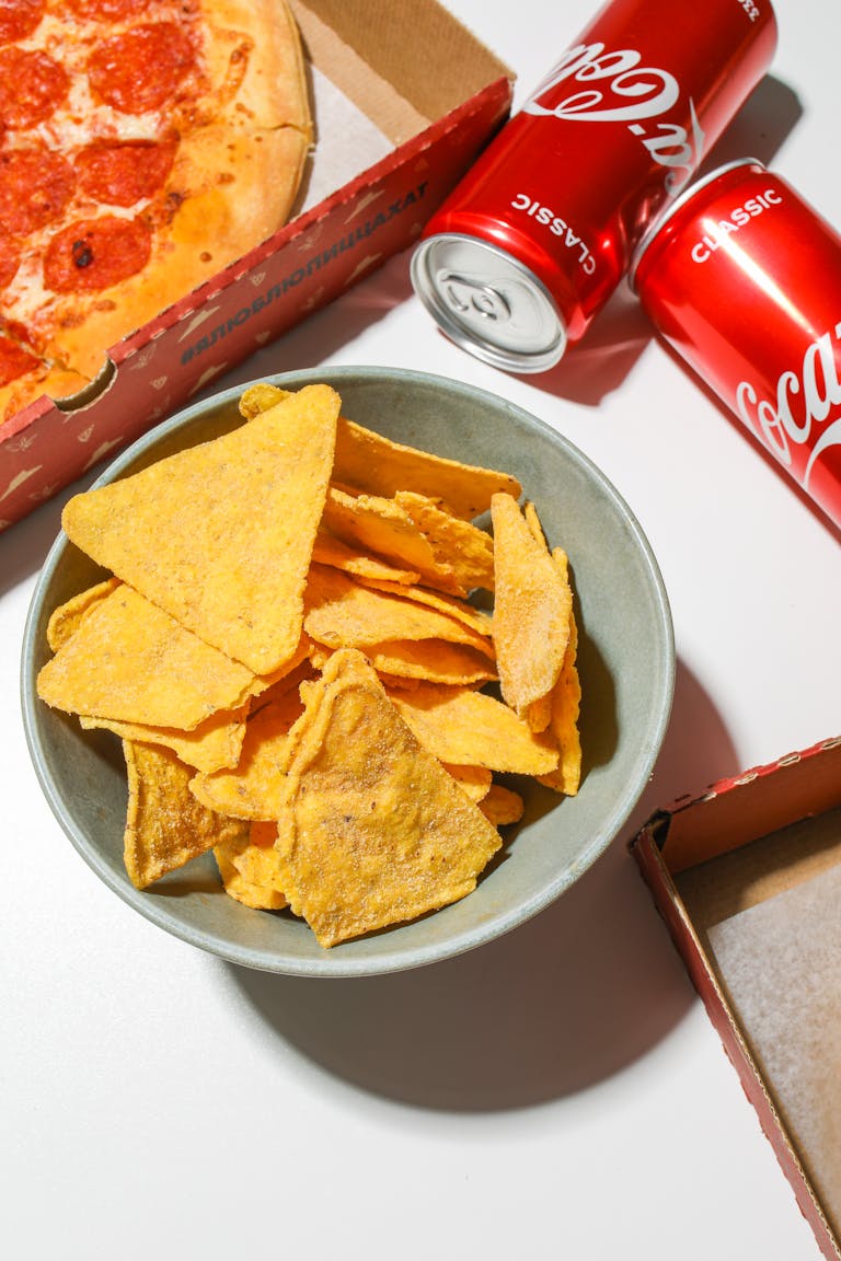 A bowl of tortilla chips with pizza and cola cans, perfect for a party snack.