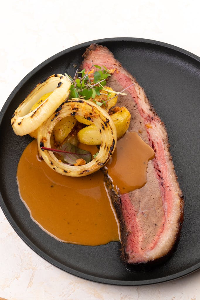 Delicious grilled pork with onion rings, potatoes, and sauce served on a black plate.