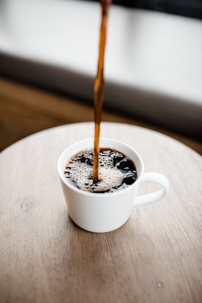 Steaming black coffee being poured into a white ceramic mug on a wooden table.