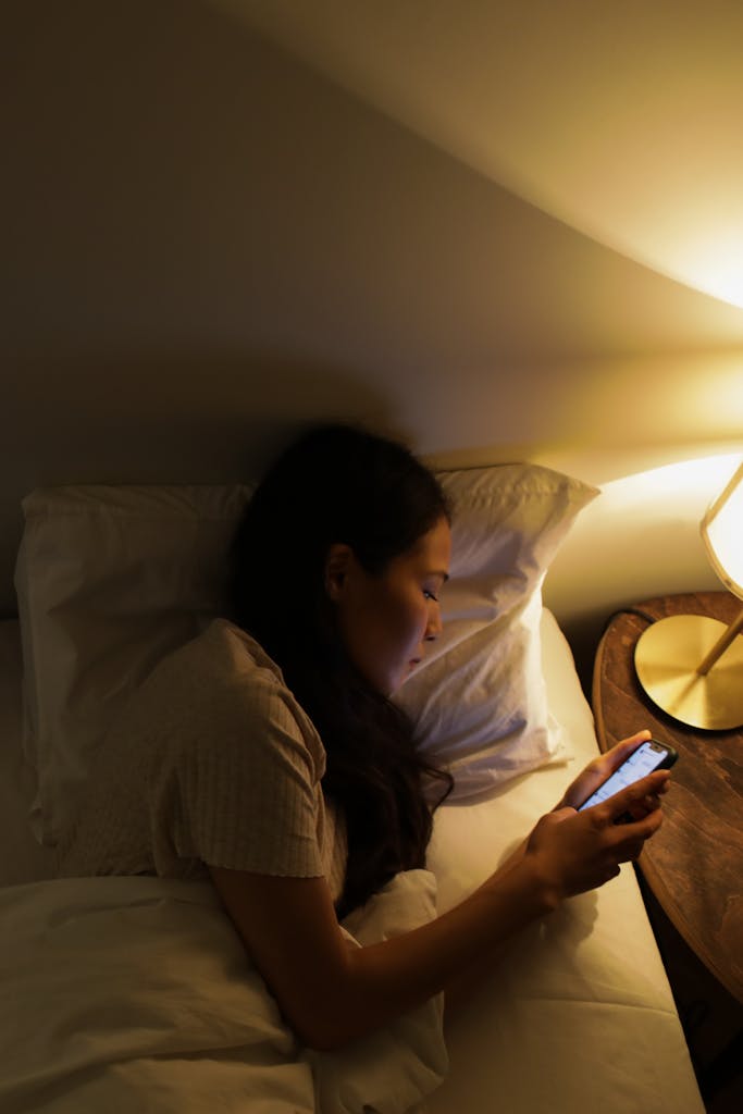 An Asian woman lying in bed under warm light using a smartphone.