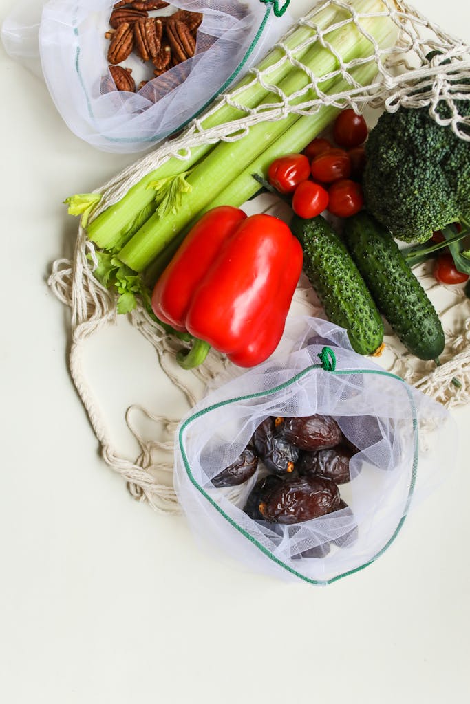 Flat lay of fresh vegetables and nuts in reusable mesh bags on a light background.