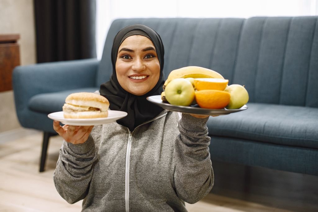 A smiling woman wearing a hijab holds a burger and a plate of fruits in a cozy indoor setting.
